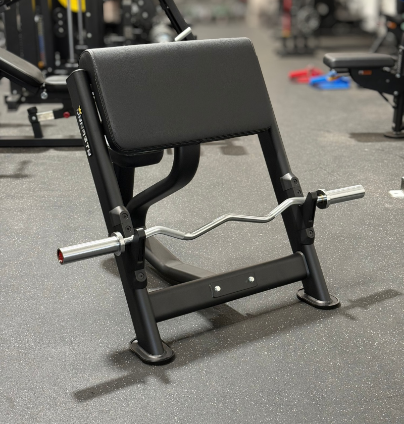 A black preacher bench with a curl barbell, photographed in a gym setting with other exercise equipment blurred in the background.