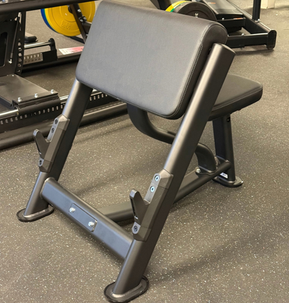 A black preacher curl bench with plastic coated bar rests on a speckled floor.