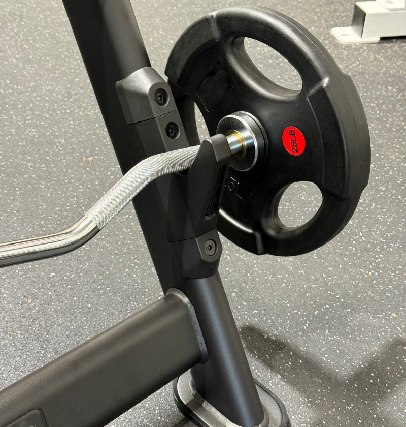 Close-up of a black weight plate on a preacher bench with a silver curved handle, set on a speckled gray gym floor.