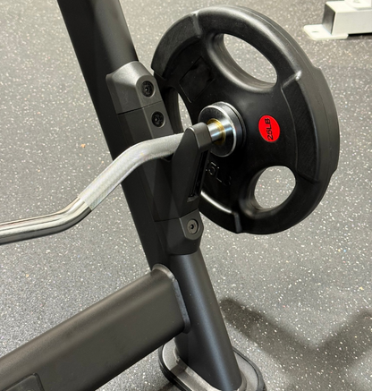 Close-up of a black weight plate on a preacher bench with a silver curved handle, set on a speckled gray gym floor.
