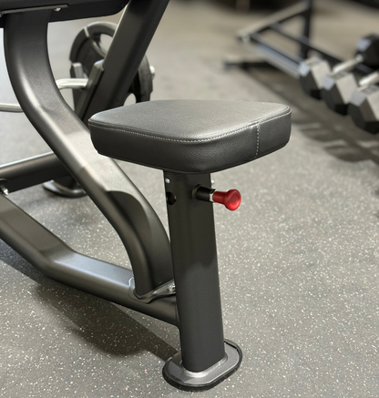 Close-up of a preacher bench seat with dark gray padded upholstery and a small red adjustment knob, set on a speckled gray gym floor.