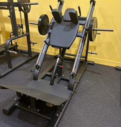 A black hack squat machine, set on a dark rubber gym floor against a yellow wall.