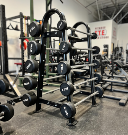 A black and silver fixed straight barbell with marked weight plates displayed on a black metal rack in a gym setting