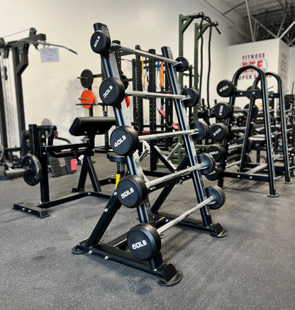 A black and silver fixed straight barbell with marked weight plates displayed on a black metal rack in a gym setting